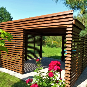 Altanka model Sewilla na tle ogrodu z kwiatami. Altanka posiada dwa wejścia i jest zabudowana poziomymi listewkami.